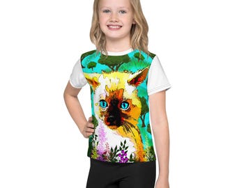 Puede incluir: Una camiseta blanca de manga corta con una colorida ilustración de un gato siamés con brillantes ojos azules. El gato está sobre un fondo turquesa con árboles y flores. La camiseta la lleva una persona joven.
