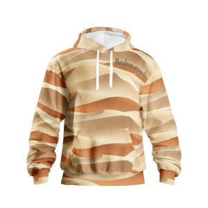 May include: A hooded sweatshirt with a desert landscape pattern in shades of tan, brown, and cream. The hoodie features a front pocket and white drawstrings. The words "Broken goods" are printed across the chest.