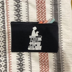May include: Black canvas zippered pouch with a white silhouette of a person reading a book on top of a stack of books. The text on the pouch reads "A reader lives a thousand lives before they die."