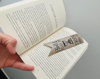May include: A brown leather bookmark with a black hourglass design and the text "So Many Books, So Little Time!" The bookmark is placed in an open book.