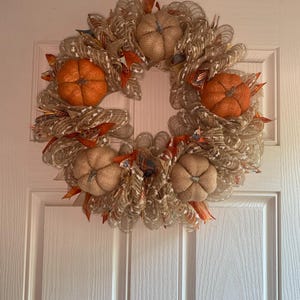 May include: A fall-themed wreath with a circular design. The wreath features five burlap pumpkins in shades of orange and tan, interspersed with decorative ribbons and leaves. The wreath is displayed on a white door.