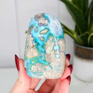 May include: A polished stone with a mix of blue, turquoise, and beige colors. The stone has a rounded top and a textured surface. The stone is being held in a hand with red nail polish.