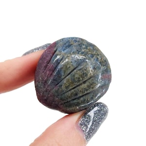 May include: A carved gemstone in shades of blue, green, and red is held between two fingers. The stone has a rounded shape with carved lines on the surface. The gemstone is a natural stone.