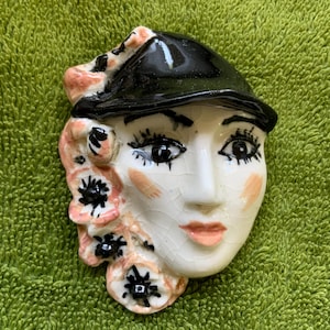 May include: A ceramic face with a black hat, pink flowers, and a white face with painted features. The face has black eyelashes, eyebrows, and a pink blush. The background is a green, textured surface.