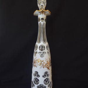 May include: A tall, white glass decanter with a decorative stopper. The decanter features gold and brown floral designs and cut-out patterns. The stopper and neck have gold accents. The decanter is approximately 14 inches tall.