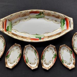 May include: A white oval serving platter with handles, accompanied by six smaller oval dishes. The set features a gold rim and decorative accents of red, green, and gold. The pieces are arranged on a dark surface.