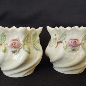 May include: Two cream-colored ceramic vases with a ruffled rim and a swirling design. Each vase features a pink rose, green leaves, and small blue flowers. The vases are decorative.