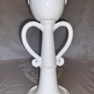May include: A tall, white ceramic vessel featuring a wide, bowl-shaped top with a teardrop-shaped relief detail. The central column is flanked by two symmetrical, curved handles that loop outward and inward. The base is circular with a beaded edge.