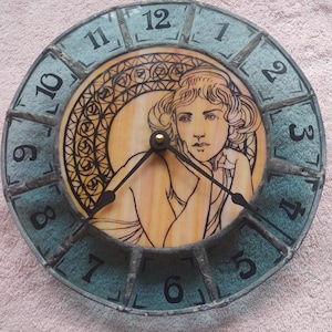 May include: Round wall clock with a teal and black frame. The clock face features a woman's portrait in warm tones, surrounded by a decorative border. Black numerals mark the hours. The clock hands are black.