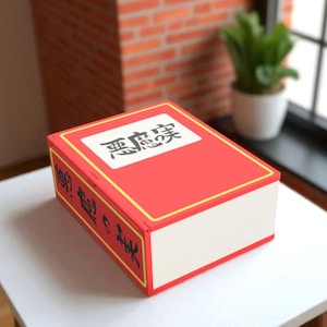 May include: A red rectangular box, designed to look like a book, with white and yellow trim. Japanese text is displayed on the top and side. The box is on a white surface. The background includes a brick wall and a plant.