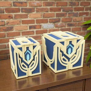 Op de afbeelding: Twee decoratieve blauwe en crèmekleurige dozen met ingewikkelde geometrische ontwerpen. De dozen hebben een scharnierend deksel en een centraal lichtblauw accent. Ze worden getoond op een houten oppervlak tegen een bakstenen muur.