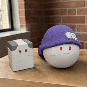 May include: A white and gray box with red dots and a white, round-shaped object with a purple hat and red dots. The objects are on a wooden surface with a brick wall background.