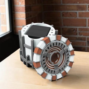 May include: A cylindrical white and gray container with a removable circular lid featuring a copper-colored segmented rim and a central mesh detail. The main body has a vertical grid structure and a black square solar panel on the top surface.