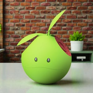 May include: A green, pear-shaped electronic device with two oval-shaped eyes and a small red section. Two green leaf-like structures extend from the top. The device is on a white surface, with a brick wall in the background.