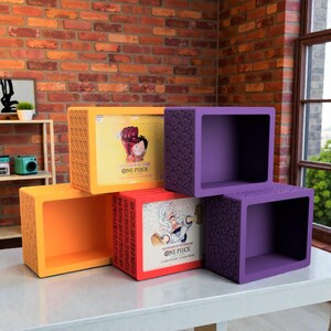 May include: A stack of four square, open-faced storage cubes in shades of orange and purple. Two cubes are orange, one with a patterned design. The other two are purple, one with a patterned design. The cubes have images from the anime series "One Piece".
