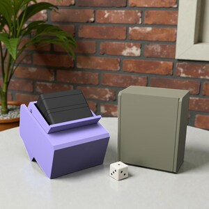 May include: A light purple angled card holder containing a stack of black cards, a solid olive green rectangular storage box, and a single white six-sided die with black pips, all arranged together on a smooth surface.