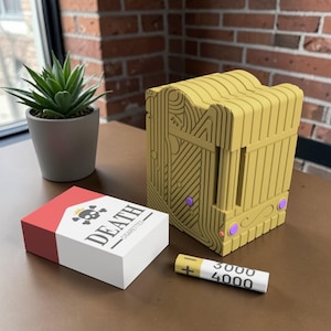 May include: A yellow, decorative box with intricate line designs and purple accents, next to a pack of "DEATH CIGARETTES" with a skull and crossbones. A small battery with "3000 4000" printed on it is also present.