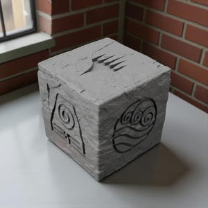 Könnte beinhalten: Eine graue, würfelförmige Skulptur mit eingravierten Symbolen, die Luft, Wasser, Erde und Feuer darstellen. Der Würfel hat eine strukturierte Oberfläche und wird auf einer weißen Oberfläche in der Nähe eines Fensters und einer Backsteinmauer ausgestellt.