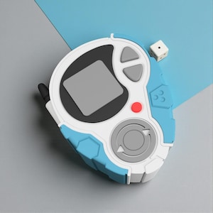May include: A blue, white, and gray handheld device with a small screen, buttons, and a circular control. A small white die with black dots is next to the device. The device has a retro, futuristic design.