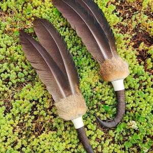 Könnte beinhalten: Zwei braune Federfächer mit weißen Spitzen und braunem Fell an der Basis der Federn. Die Fächer sind an braunen, gebogenen Griffen befestigt.