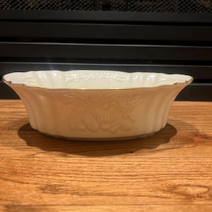May include: Off-white oval serving bowl with a gold rim and embossed floral design. The bowl has a scalloped edge and is made of ceramic. A decorative piece for serving food or displaying items.
