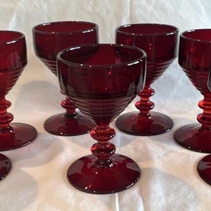 May include: A set of six deep red glass goblets, each featuring a wide bowl with horizontal ribbed detailing, a slender stem with two spherical nodes, and a circular base. The glassware is uniform in color and design, showcasing a translucent finish.