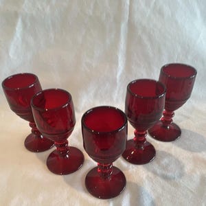 May include: A set of five matching ruby red glass goblets, each featuring a rounded bowl, a short stem with a decorative ringed detail, and a circular base. The glassware has a deep, translucent crimson hue and a smooth, polished finish.