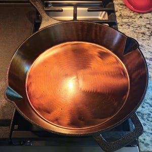 May include: A seasoned cast iron skillet with a copper-coloured interior sits on a gas hob. The skillet has a handle and a rounded shape, ready for cooking. A red bowl is visible in the background.