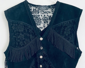 May include: Black suede vest with lace and fringe detailing. The vest features a V-neck, decorative silver buttons, and intricate floral lace panels. The bottom and sides of the vest are adorned with long fringe.
