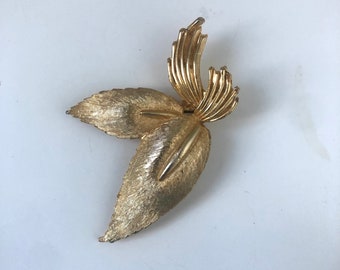 May include: Gold-toned leaf brooch with a textured finish. The brooch features two large leaves and a cluster of smaller, stylized leaves at the top. The design is symmetrical and detailed, with visible veining on the leaves.
