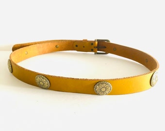 Peut inclure: Ceinture en cuir jaune moutarde ornée de conchos floraux argentés décoratifs. La boucle rectangulaire et les passants sont en laiton. Un accessoire classique pour jeans ou jupes.