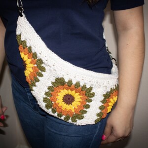 May include: A white crocheted bag with a sunflower design. The bag has a long strap and is worn across the body.