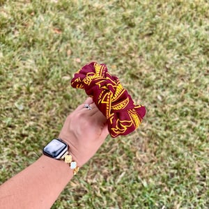 May include: A maroon fabric scrunchie featuring a repeating yellow graphic pattern and the text "COMMUNITY" printed in a bold, sans-serif font. The accessory has a gathered, ruffled texture and is constructed from a soft, flexible material.