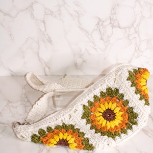 May include: A white crocheted bag with a sunflower design. The bag has a long strap and is made with green, yellow, orange, and brown yarn.