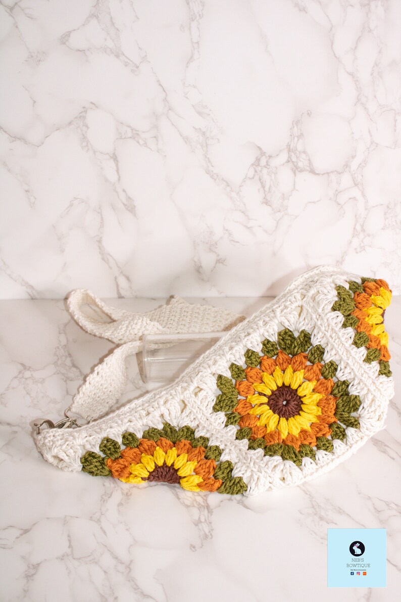 May include: A white crocheted bag with a sunflower design. The bag has a long strap and is made with green, yellow, orange, and brown yarn.