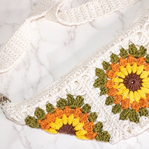May include: A white crocheted bag with a long strap and a sunflower design. The bag is made with different colored yarn, including green, orange, and yellow.