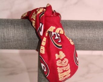 Puede incluir: Diadema de tela roja con el logotipo y el texto de los 49ers de San Francisco en dorado y blanco. La diadema está anudada y se muestra en un soporte gris. Un accesorio para el cabello.