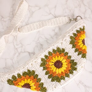 May include: A white crocheted fanny pack with a long white strap. The fanny pack features three crocheted sunflowers with brown centers, yellow petals, orange accents, and green leaves.