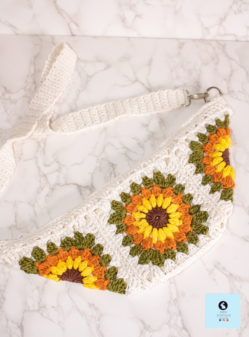 May include: A white crocheted fanny pack with a long white strap. The fanny pack features three crocheted sunflowers with brown centers, yellow petals, orange accents, and green leaves.