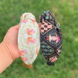 May include: Two fabric headbands with knotted ends. One headband is green with a white and pink floral pattern featuring birds. The other headband is black with a colorful pattern featuring a woman's face and geometric shapes.