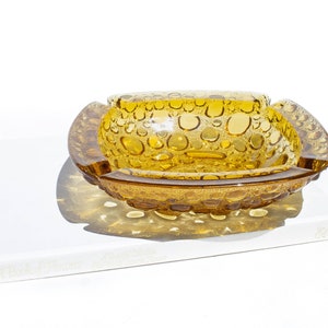 Peut inclure: Un cendrier vintage en verre ambré avec un motif texturé en bulles. Le cendrier est de forme ovale avec un bord surélevé et repose sur une surface blanche.