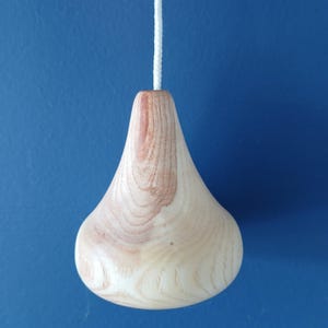 Puede incluir: Un tirador de luz de madera de color claro con un cuerpo liso en forma de gota y patrones de veta natural visibles. El objeto est&aacute; unido a un cord&oacute;n trenzado blanco que se extiende verticalmente desde el centro superior de la pieza de madera.