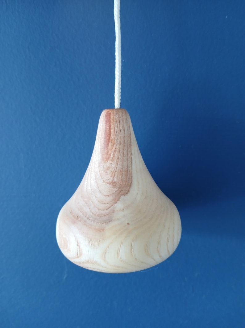 Puede incluir: Un tirador de luz de madera de color claro con un cuerpo liso en forma de gota y patrones de veta natural visibles. El objeto est&aacute; unido a un cord&oacute;n trenzado blanco que se extiende verticalmente desde el centro superior de la pieza de madera.