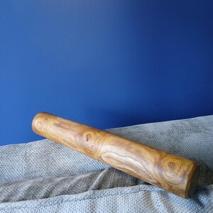 Può includere: Un mattarello cilindrico in legno con un motivo a venature naturali. Il mattarello è marrone chiaro con nodi e vortici più scuri. Poggia su una superficie grigia strutturata contro uno sfondo blu.