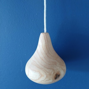 Puede incluir: Un tirador de luz de madera clara con un cuerpo liso en forma de l&aacute;grima y patrones de veta de madera natural visibles. El objeto presenta un peque&ntilde;o nudo oscuro en el lado inferior derecho y est&aacute; unido a un cord&oacute;n blanco trenzado en la parte superior.