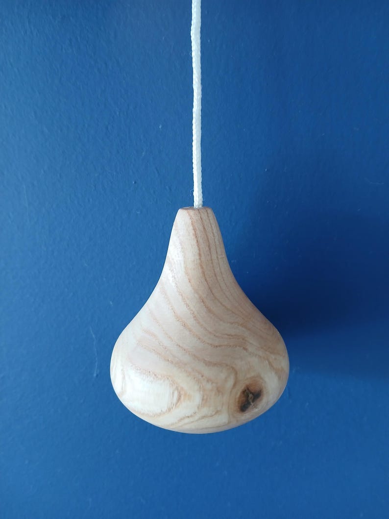 Puede incluir: Un tirador de luz de madera clara con un cuerpo liso en forma de l&aacute;grima y patrones de veta de madera natural visibles. El objeto presenta un peque&ntilde;o nudo oscuro en el lado inferior derecho y est&aacute; unido a un cord&oacute;n blanco trenzado en la parte superior.