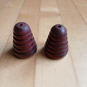 Puede incluir: Dos objetos de madera marrón oscuro en forma de cono, con una serie de ranuras horizontales. Cada objeto tiene un pequeño agujero en la parte superior. Los objetos se muestran sobre una superficie de madera marrón claro.