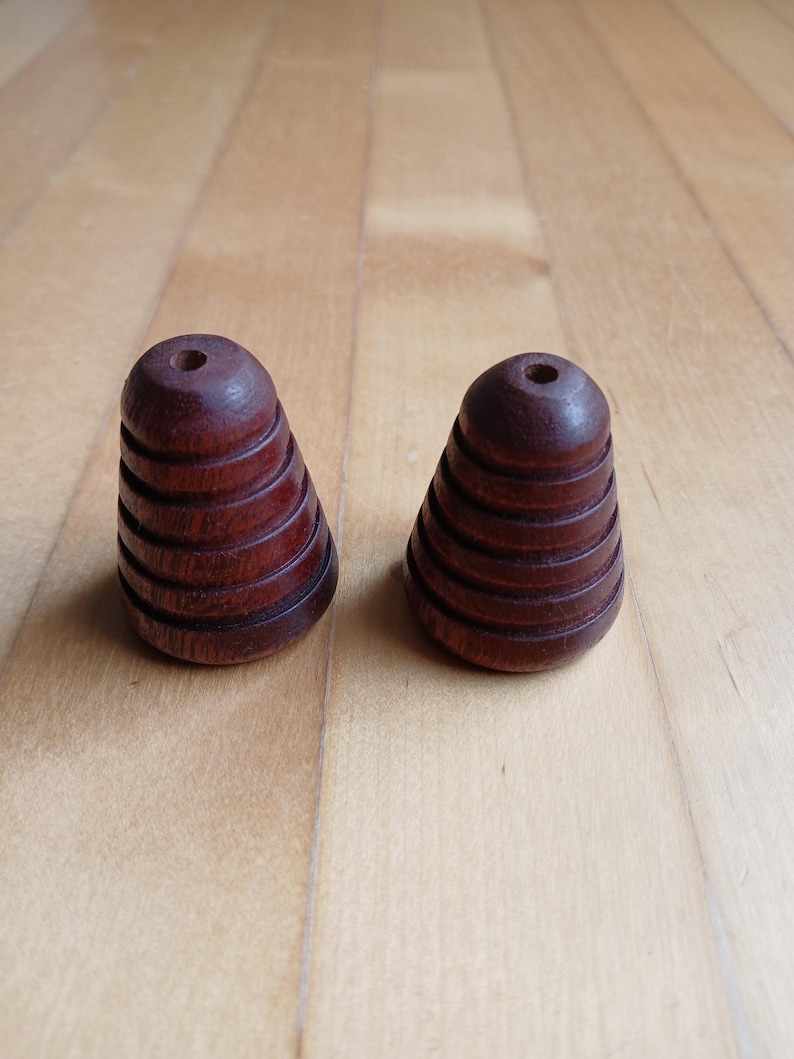 Peut inclure: Deux objets en bois brun fonc&eacute; en forme de c&ocirc;ne, avec une s&eacute;rie de rainures horizontales. Chaque objet a un petit trou en haut. Les objets sont pr&eacute;sent&eacute;s sur une surface en bois brun clair.