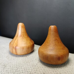 Puede incluir: Dos objetos de madera de color marr&oacute;n claro, lisos y con forma redondeada de gota. Cada pieza presenta una base circular ancha que se estrecha hacia arriba hasta una parte superior plana y estrecha con una peque&ntilde;a abertura central, mostrando las vetas naturales.