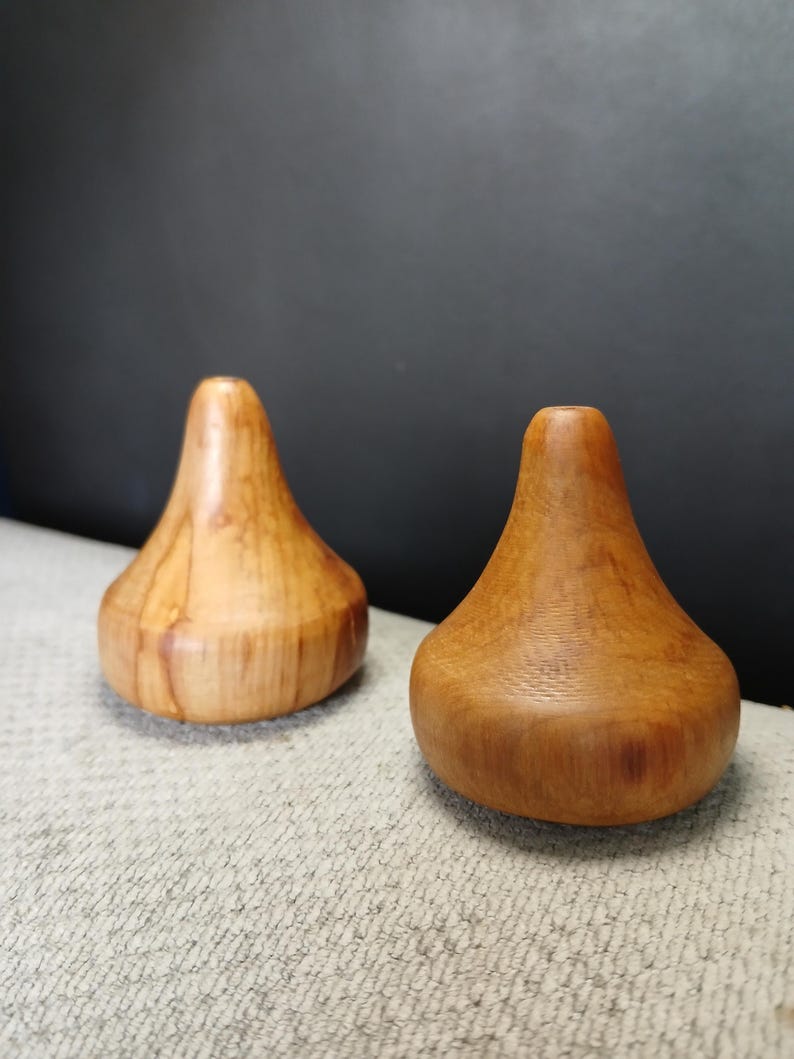 Puede incluir: Dos objetos de madera de color marr&oacute;n claro, lisos y con forma redondeada de gota. Cada pieza presenta una base circular ancha que se estrecha hacia arriba hasta una parte superior plana y estrecha con una peque&ntilde;a abertura central, mostrando las vetas naturales.